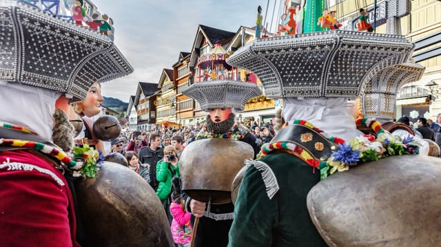 Verkleidete Gruppe mit farbigen Masken, Hauben und Glocken. Im Hintergrund Fassaden alter Häuser und Menschenmassen.