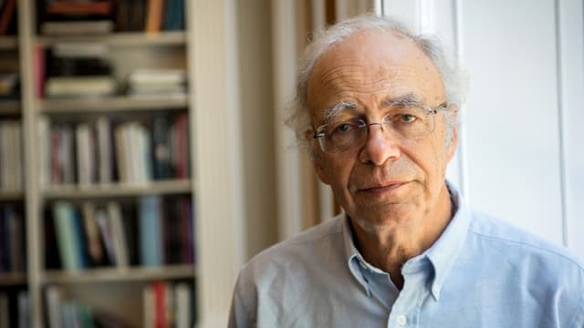 Peter Singer, vor einem Büchergestell stehend, gerade in die Kamera blickend.