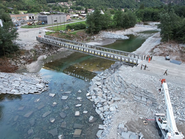 Eine metallbrücke über einem Fluss, Menschen stehen darum herum, einzelne Baugerätschaften stehen 10 Meter daneben.