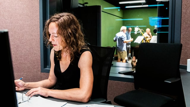 Ein Frau sitzt in einem Raum vor einem Zettel. Hinter ihr sieht man durch ein Fenster in ein Radiostudio.