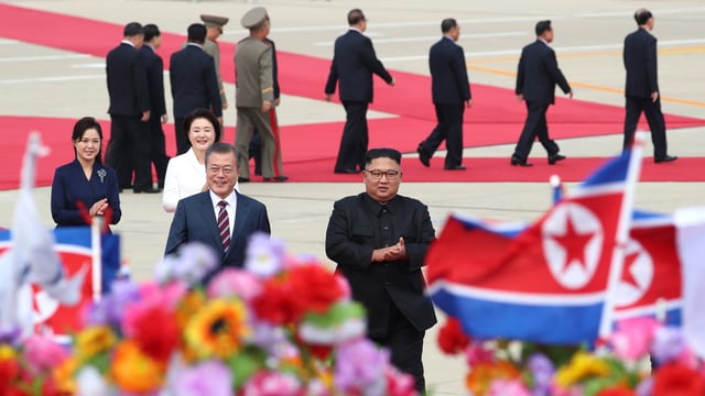 Moon und Kim mit ihren Frauen auf rotem Teppich, im Vordergrund Fahnen und Blumen.