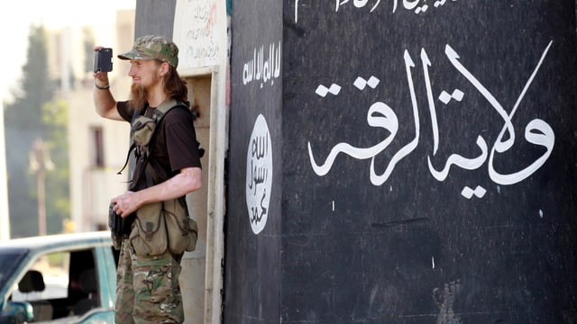 Aufnahme aus Rakka: Bewaffneter Mann mit rot-blondem Bart vor einer IS-Flagge mit Filmkamera in der Hand.