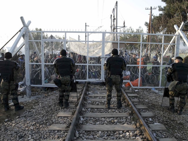 Polizisten in Kampfmontur stehen vor einem Gittertor, das ein Bahngeleise geschlossen hält, dahinter viele Menschen.