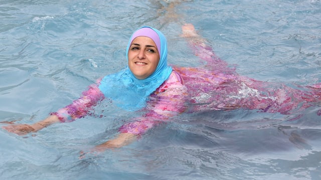 Eine Frau badet in einem rosa Burkini mit helllblauem Kopftuch.