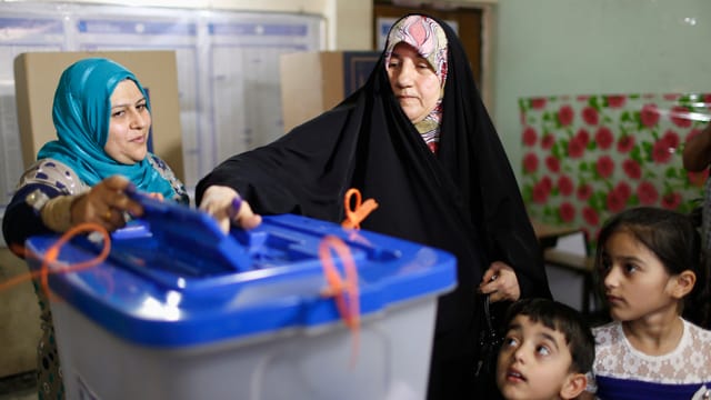 Eine Frau wirft ihren Wahlzettel in die Urne, daneben steht eine Wahlhelferin, an der Seite der Wählerin zwei Kinder.