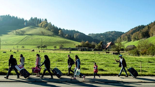 Acht Personen mit Koffern auf einem Trottoir, im Hintergrund grüne Wiesen und Hügel.