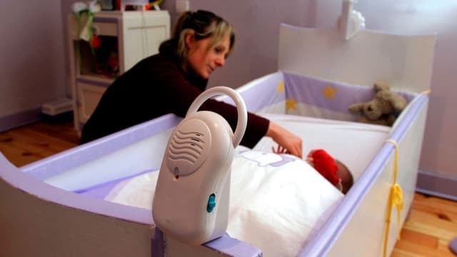 Mutter bettet ihr Kleinkind in die Wiege, im Vorgergrund steht ein Babyphone.