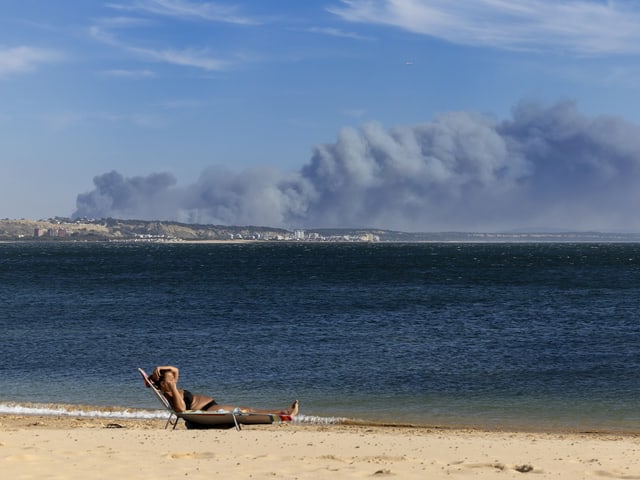 Person auf Sonnenliege am Strand mit Rauchwolken in der Ferne.