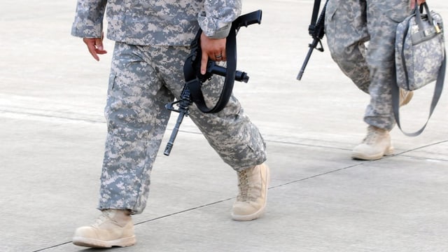 Zwei US-Soldaten (nur die Beine), gehend, in Uniform, mit einem Maschinengewehr in der Hand.