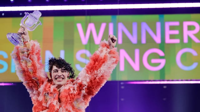 Person in orangefarbenem Federkostüm hält Pokal hoch und jubelt vor 'WINNER SINGING CONTEST'-Anzeige.