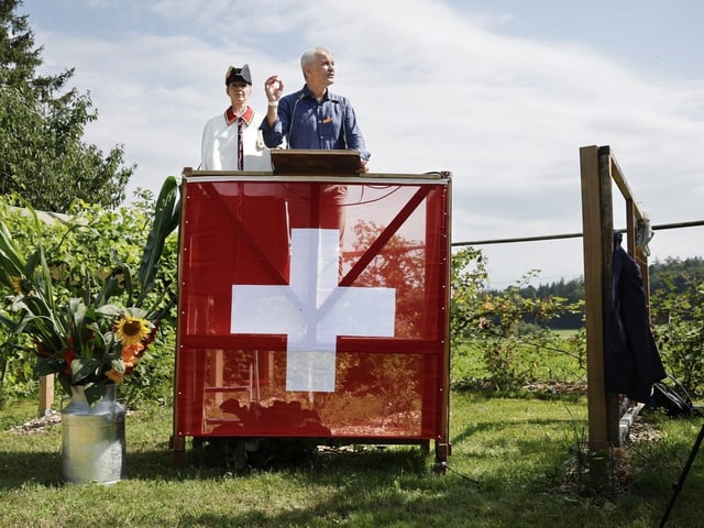 Mann hält Rede auf mit Schweizer Fahne dekoriertem Podium im Freien.