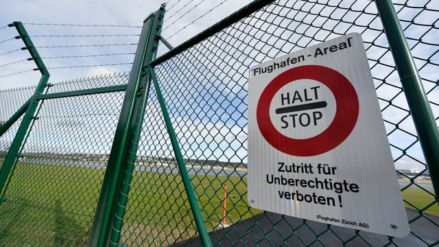 Ein Schild hängt an einem Zaun, darauf steht: "Halt - Stop". Dahinter ist eine Landebahn am Zürcher Flughafen.