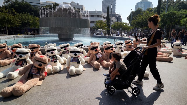 Eine Mutter und ein Kind im Kinderwagen betrachten ein Mahnmal in Tel Aviv aus Teddybären mit verbundenen Augen.