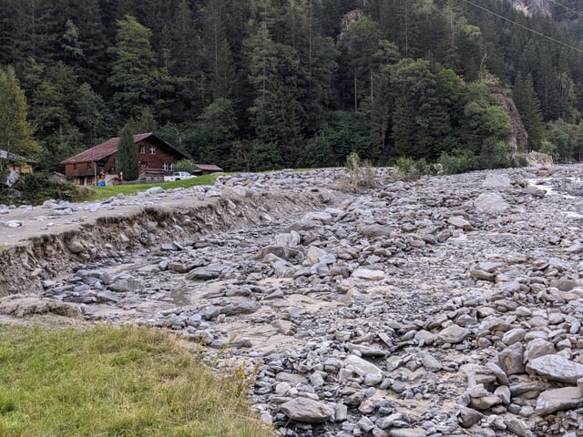 Bachlauf mit Geröll und einem Haus im Hintergrund.