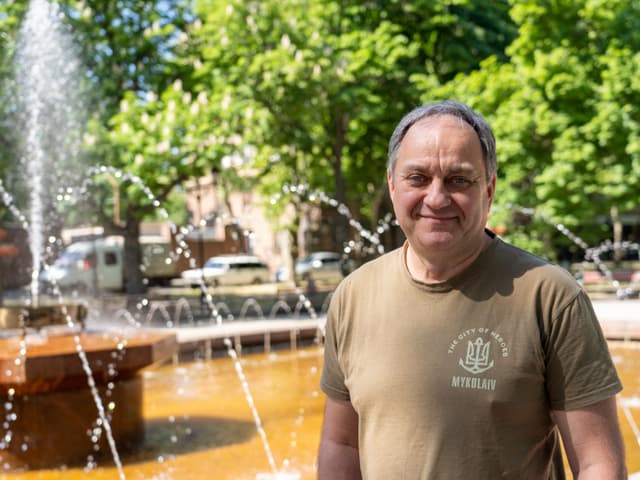 Mann lächelt vor einem sprudelnden Brunnen im Freien.