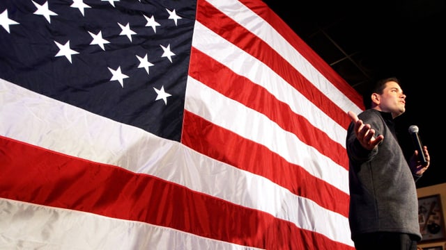 Rubio mit einem Mikrofon in der Hand vor einer riesigen US-Flagge.