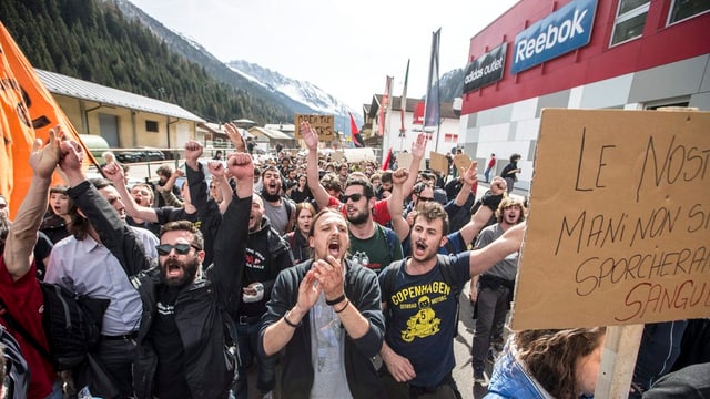 Ein Menschenumzug. Links erheben sich Berge, rechts ein Gebäude. 