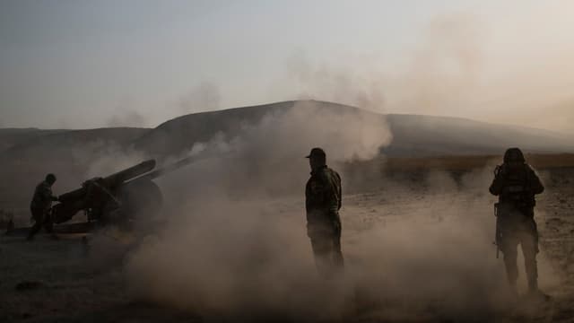 Drei kurdische Kämpfer stehen im Staub neben einer Artillerieeinheit.