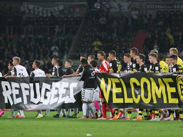 Ein grosses Banner wird von den Spielern getragen. Darauf steht: Refugee are welcome.