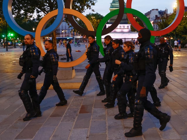 Polizisten in schwarzer Uniform vor olympischen Ringen.