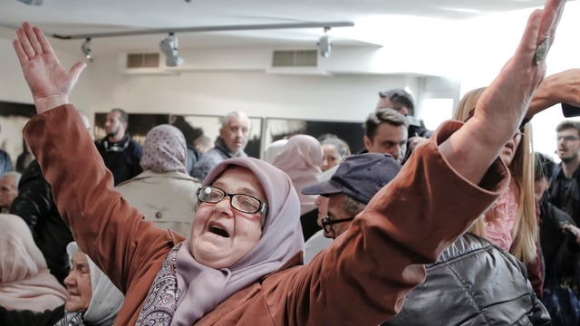 Eine Frau mit Brille und Kopftuch mit erhobenen Händen.