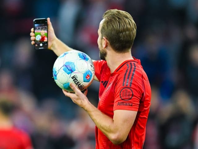 Fussballspieler in rotem Trikot nimmt Selfie mit Ball auf dem Spielfeld auf.