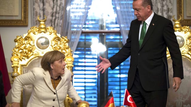 Die deutsche Bundeskanzlerin Angela Merkel wird sitzend überrascht vom Händedruck des türkischen Staatschefs Recep Tayyip Erdogan.