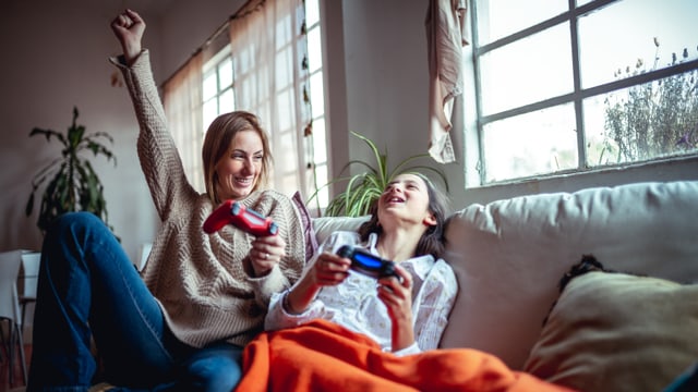 Eine Mutter spielt mit ihrer jugendlichen Tochter Videospiele. Sie sitzen auf der Couch und haben Controller in der Hand