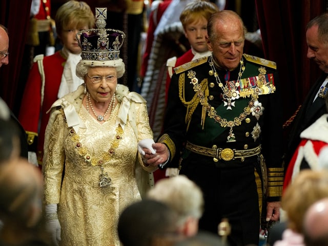 The old lady with a crown holds the hand of an elderly gentleman in a suit and tie.