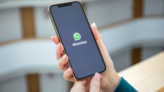 Zwei Hände halten Smartphone, Auf Bildschirm grünes Whatsapp-Logo, schwarz hinterlegt