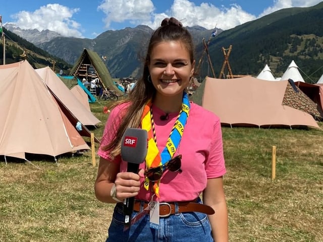 Angela mit dem SRF Mikrofon in der Hand. Im Hintergrund sieht man eine Wiese mit Pfadizelten.