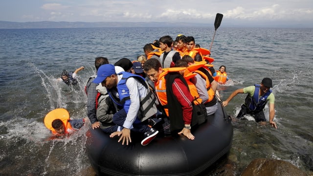 Ein überfülltes Gummiboot landet an, an Bord Dutzende Personen, sie tragen Schwimmwesten.