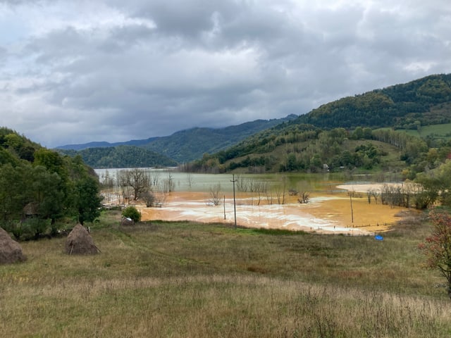 Der See besteht aus orangem und grauem Wasser. Im Vordergrund eine grün-braune Wiese.