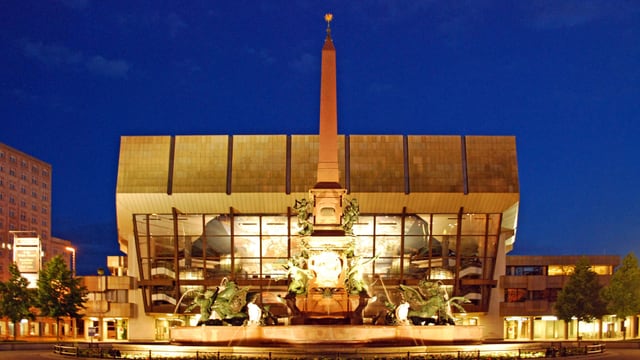 Ein grosser Brunnen mit Turm in der Mitte. Im hintergund ein rechteckiges, modernes Gebäude. Nacht.