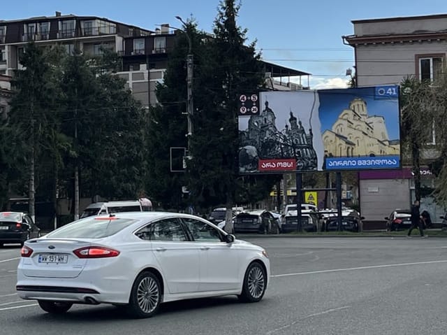 Weisse Limousine vor Gebäude mit Plakatwand auf Strasse.