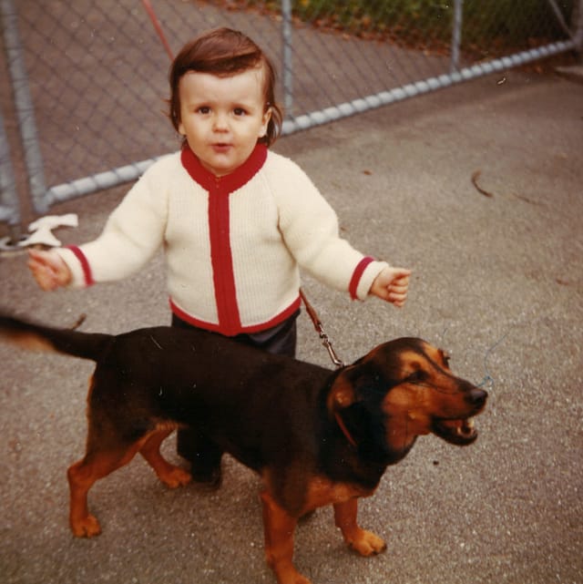 Ein Kleinkind mit rot-weisser Strickjacke steht neben einem kleinen Hund.