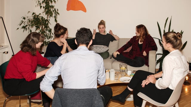 Fünf junge Frauen und ein Mann sitzen auf Stühlen und einem Sofa.