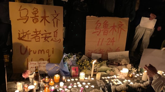 Kerzen und Blumen liegen auf der Strasse. Dahinter stehen Menschen mit Plakaten und weissen Zettel. 