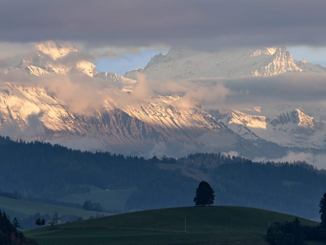 Verschneite Berge hinter Wolken mit grüner Landschaft.