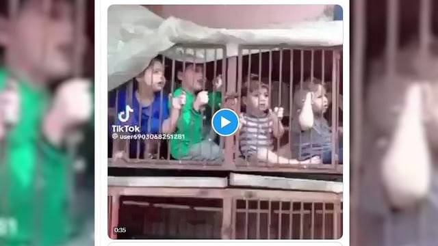 Video-Still von vier kleinen Kindern in einem rostigen Käfig, halten teils die Gitterstäbe, schauen nach draussen.