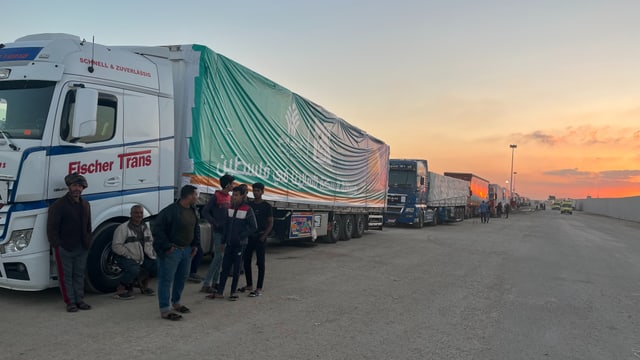 Männer stehen vor einem Lastwagen auf einer Strasse. Hinter dem Lastwagen reihen sich weitere Lastwagen bis zum Horizont