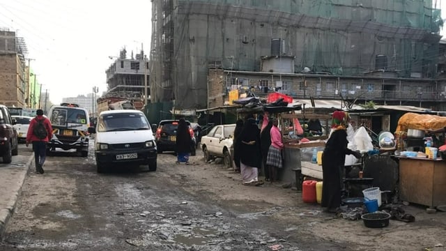 Schmutzige Strasse mit einem weissen Van, links geparkte Autos, rechts Marktstände