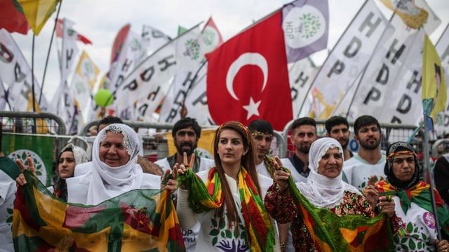 Eine Reihe Frauen und Männer in farbigen Kleidern, dazu eine türkische und viele weisse HDP-Fahnen.