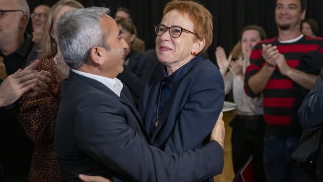 Links Mann mit grauen Haaren seitlich, rechts Frau mit kurzen, roten Haaren, umarmen sich, lachen.