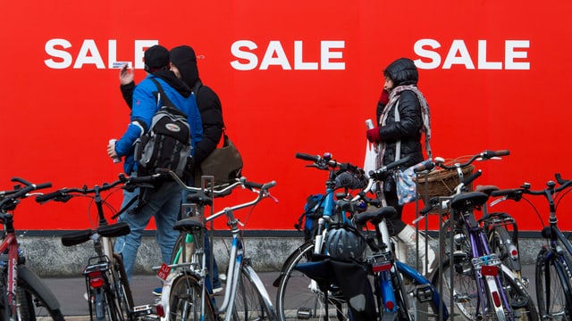 Symbolbild: Plakatwand mit «Sale»-Slogans, davor parkierte Fahhräder und einige Personen.