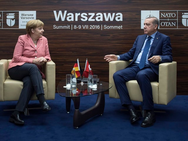 Merkel (links) und Erdoga sitzen auf schweren Stühlen und sprechen zueinander. Auf einem kleinen Tisch hat es zwei Fähnchen der beiden Länder.