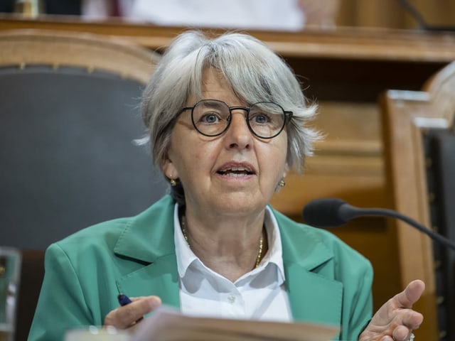 Frau mit grauen Haaren und schwarzer Brille sitzt vor einem Rednerpult in einem türkisen Blazer und weisser Bluse.