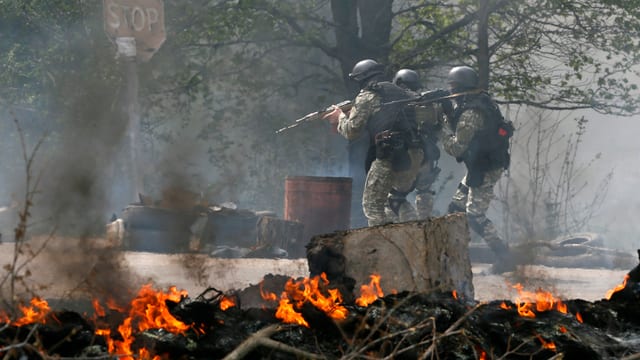 Bewaffnete Soldaten stehen vor brennenden Trümmern.