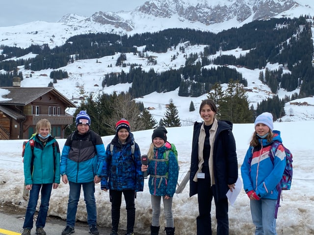 Eine Gruppe von Kindern hat sich in einer Reihe draussen aufgestellt. Hinter ihnen sieht man den Schnee und ein Holzhaus