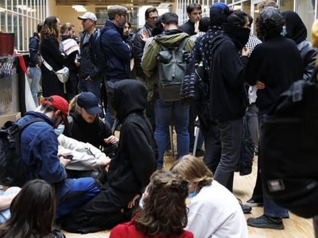 Gruppe von Menschen in einer belebten Korridorhalle, einige sitzen auf dem Boden, andere stehen.